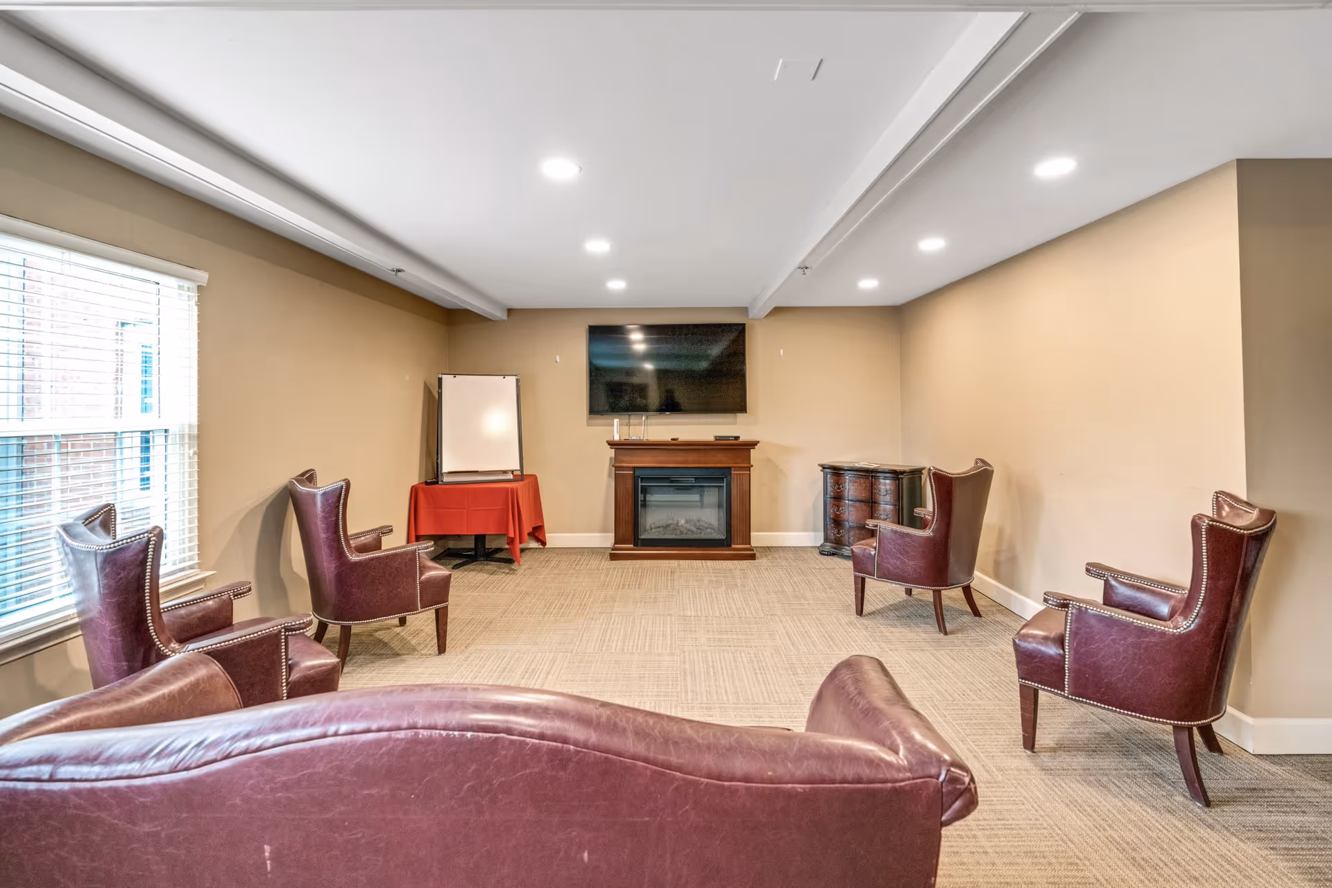 A neutral-tone common room with leather armchairs arranged facing a wall-mounted TV above an electric fireplace.