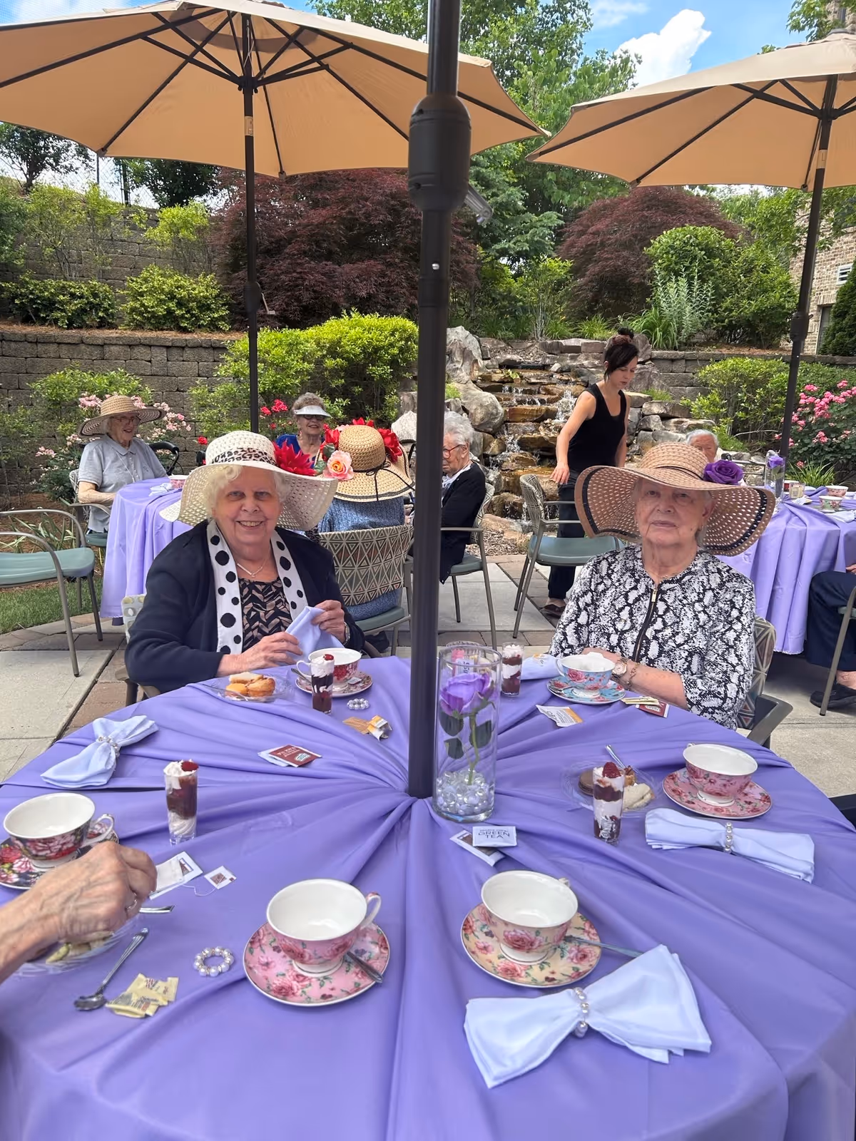 A group of elderly women wearing decorative hats sitting around a round table covered with a purple tablecloth, enjoying tea and desserts outdoors under large beige umbrellas. The setting includes a garden with green bushes, flowers, and a small waterfall feature in the background.