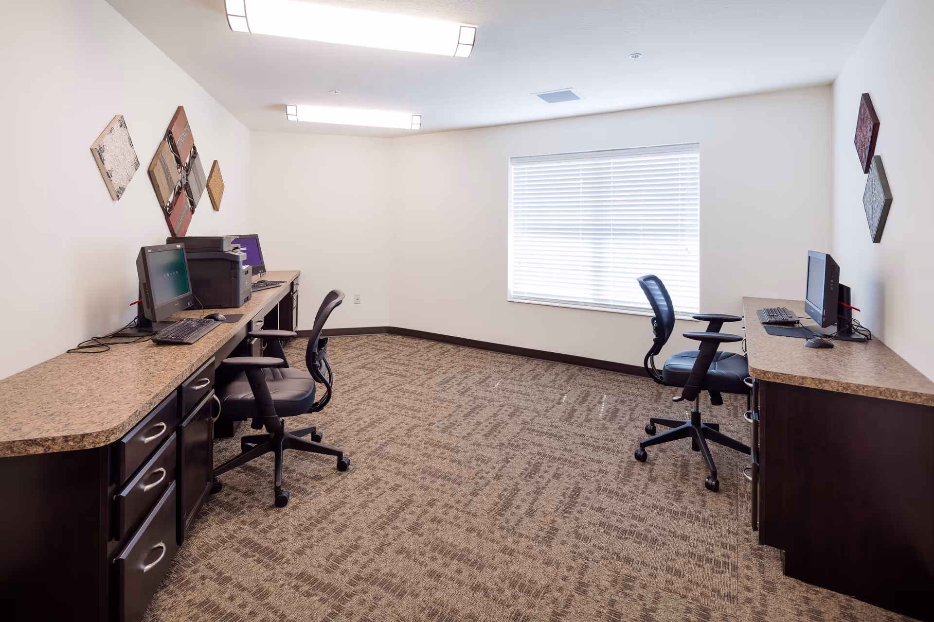 A small office room with two desks along opposite walls, each equipped with a computer and office chair. The room has beige carpet, white walls decorated with diamond-shaped wall art, and a window with closed blinds letting in natural light.