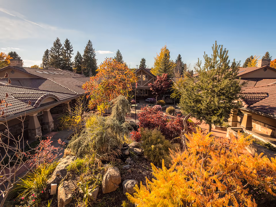 View of a senior living facility courtyard at Courtyard at Coeur d'Alene featuring a landscaped garden with various trees and shrubs in autumn colors, surrounded by buildings with brown tiled roofs under a clear blue sky.