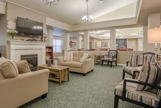 A cozy living room area in a senior living facility with beige sofas, striped armchairs, a wooden coffee table, a fireplace with a mounted flat-screen TV above it, and soft lighting from chandeliers and a floor lamp.