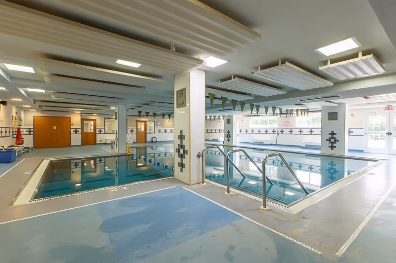 Well-lit indoor swimming pool area with handrails, tiled columns, lane flags, and poolside walkways.