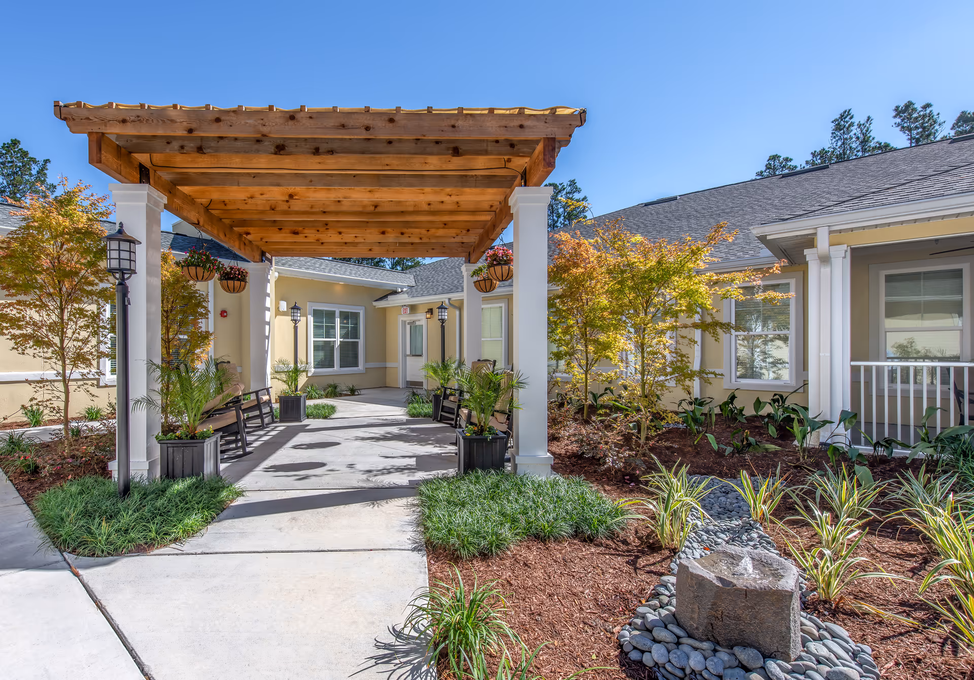 Sunny courtyard entrance featuring a wooden pergola, benches, and landscaped walkways in front of the facility.