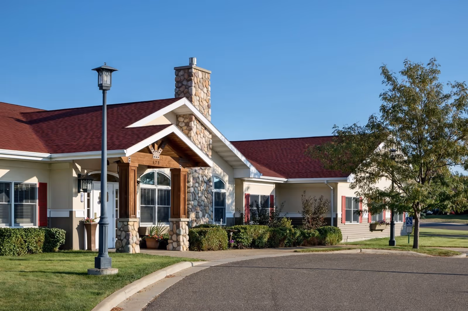 Exterior view of CountryHouse Residence building with a stone chimney, red roof, beige walls, and red window shutters. There is a lamp post and a tree near the entrance, with a curved driveway and well-maintained lawn and bushes.