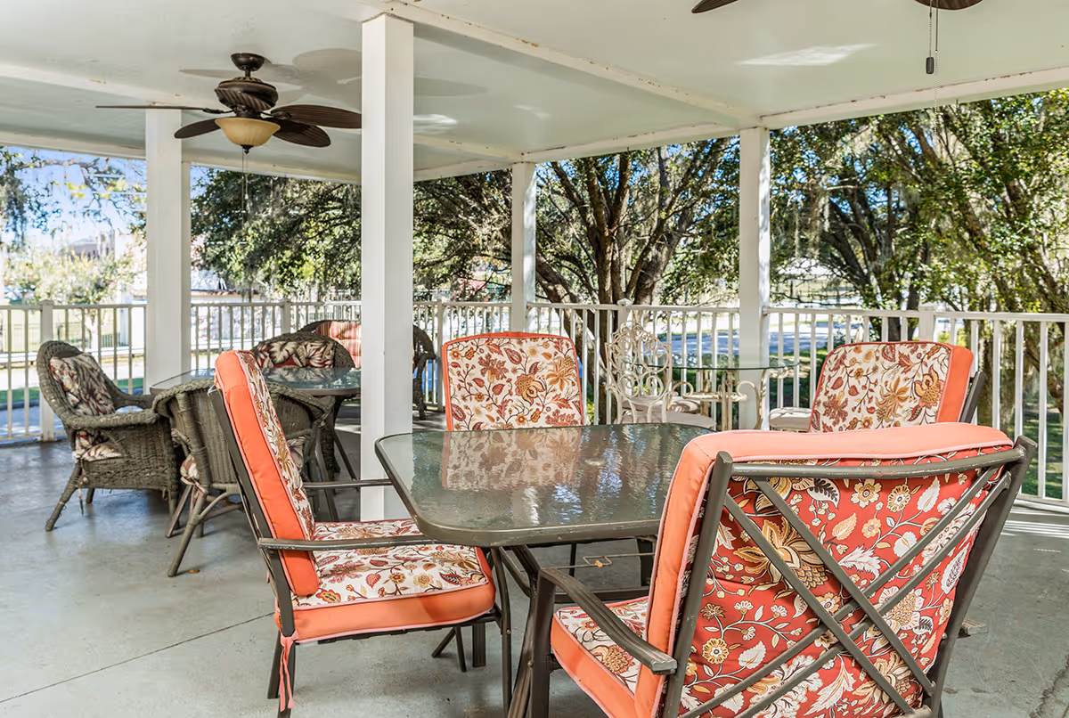 Covered outdoor patio with a glass-top table and floral-cushioned chairs under a ceiling fan overlooking trees.