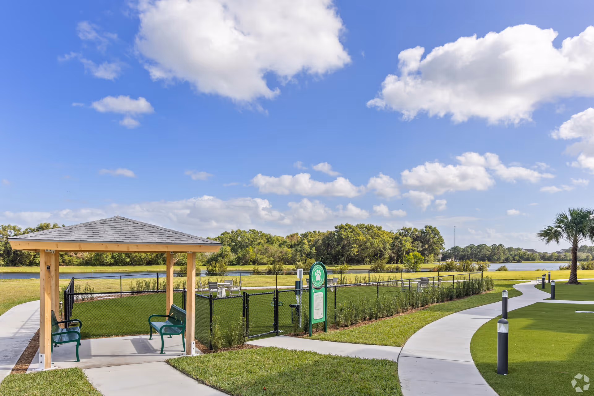 Outdoor gazebo and fenced seating area with benches beside a paved walking path overlooking a lake and landscaped grounds.