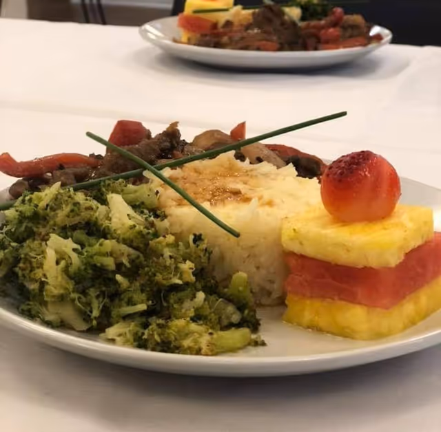A plated meal of rice, broccoli, sautéed vegetables, and stacked pineapple and watermelon topped with a cherry on a white tablecloth.