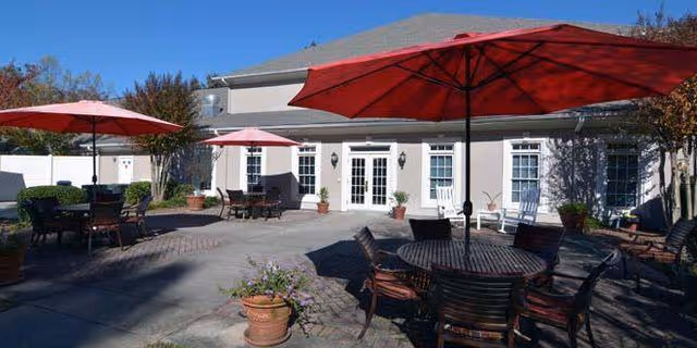 Outdoor patio area at Regency House Buford with multiple round tables and chairs under large red umbrellas, potted plants, and a building with white doors and windows in the background under a clear blue sky.