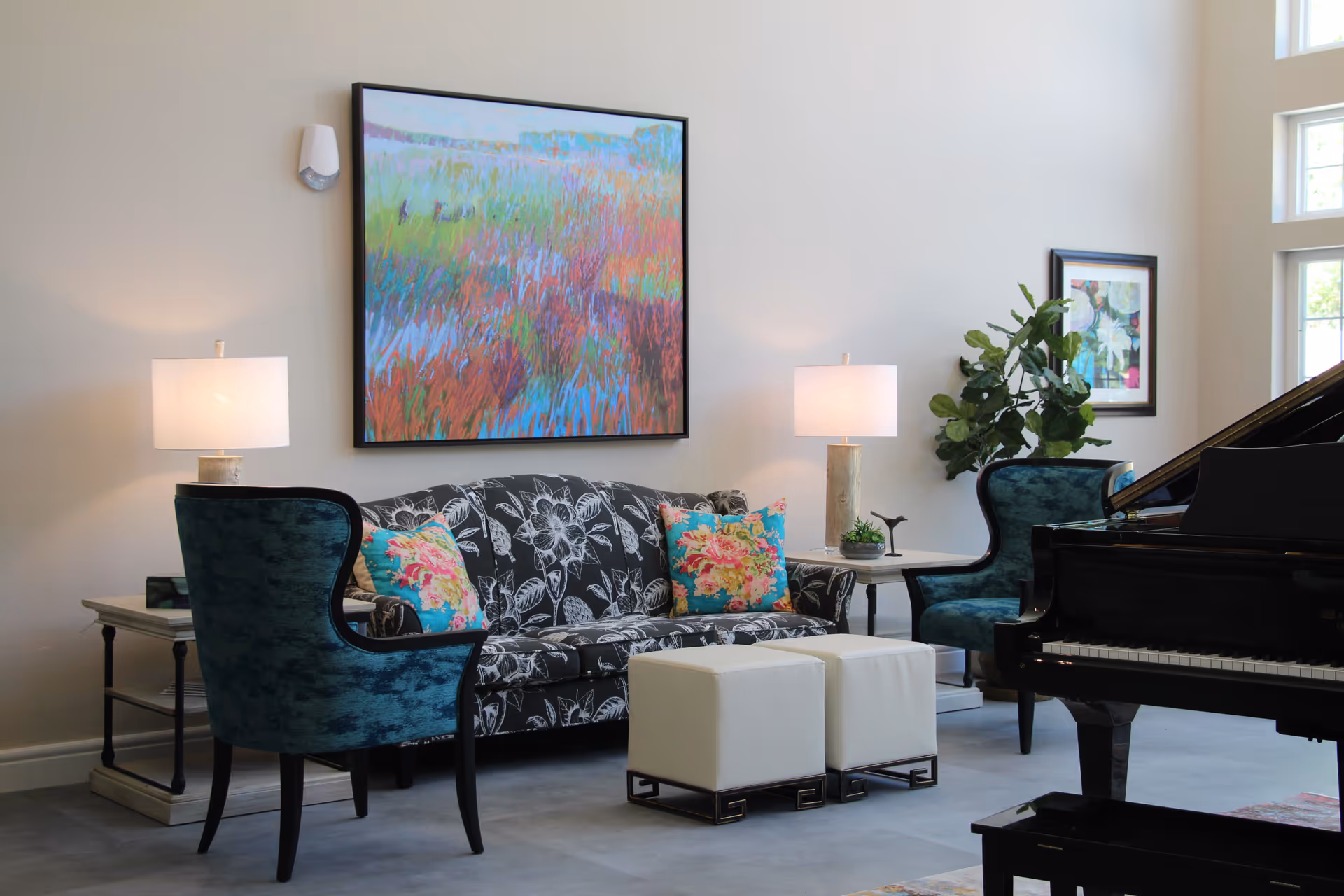 A bright and elegant living room with a black floral-patterned sofa adorned with colorful floral pillows, two teal upholstered armchairs, two white ottomans, two side tables with lamps, a large colorful abstract painting on the wall, a potted plant, and a black grand piano near tall windows.