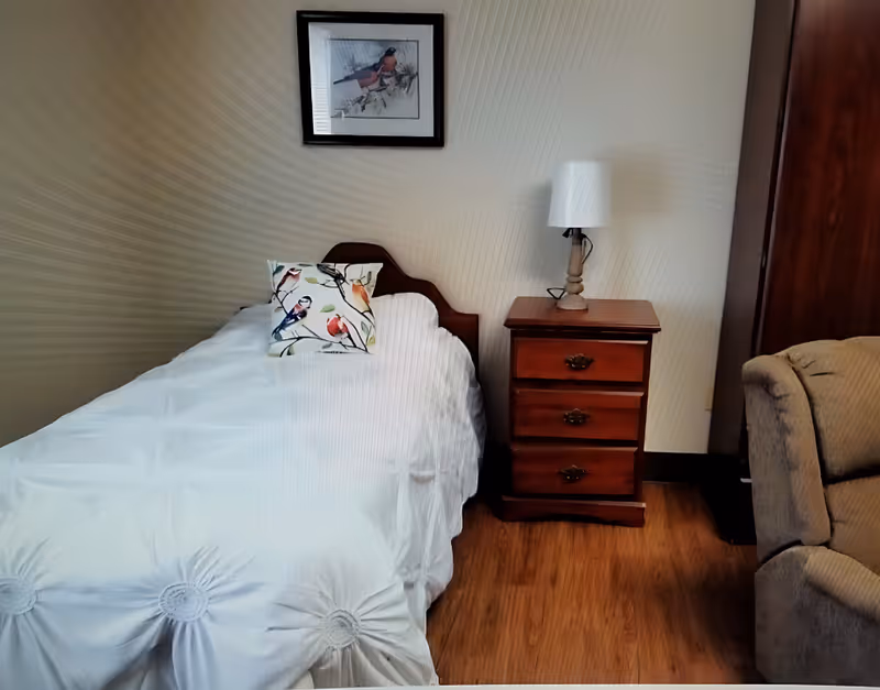 A cozy bedroom with a single bed covered in a white comforter and a decorative pillow featuring birds. Next to the bed is a wooden nightstand with three drawers and a table lamp on top. A framed picture of a bird hangs on the wall above the bed. Part of a beige upholstered chair is visible on the right side.