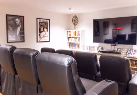 Rows of black leather recliner chairs facing a large wall-mounted TV with shelves of DVDs and movie posters on the wall.