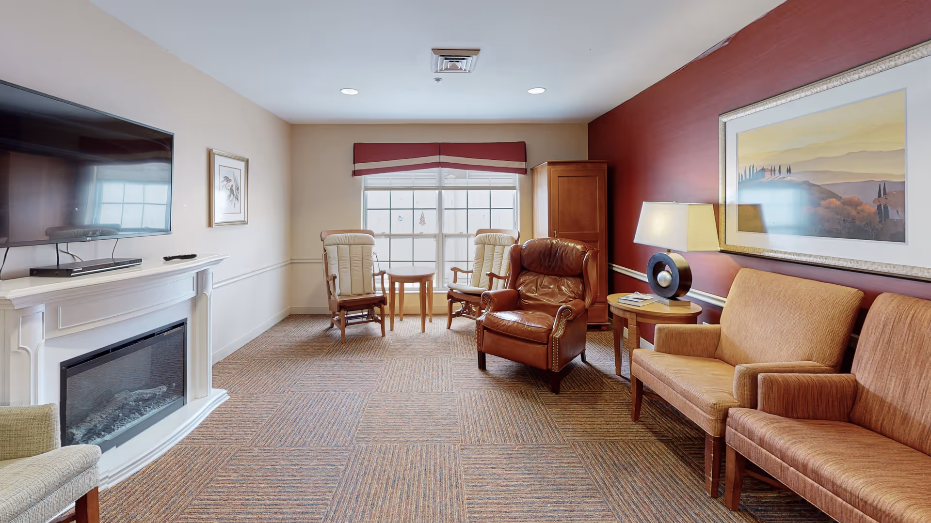 A cozy living room with a large window letting in natural light. The room features a wall-mounted flat-screen TV above a white fireplace, two beige armchairs with a small round table between them near the window, a brown leather armchair, a wooden side table with a lamp, and a beige sofa. The walls are painted beige and burgundy, with a framed landscape painting on the burgundy wall.