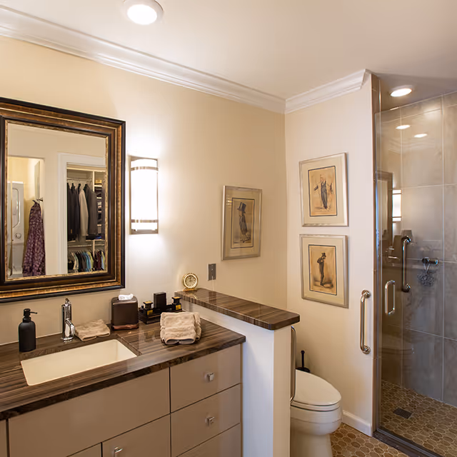 A modern bathroom with a wooden countertop and built-in sink, a large framed mirror above the sink, a wall-mounted light fixture, a toilet next to a half wall, and a glass-enclosed shower with tiled walls and floor. Two framed artworks hang on the wall above the toilet.