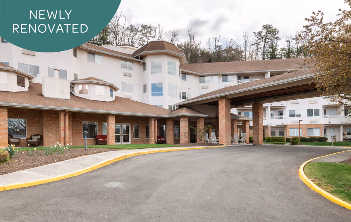 Exterior view of Holiday Elm Park Estates, a multi-story senior living facility with a covered entrance supported by brick pillars. The building has large windows and a brown roof, surrounded by a paved driveway and landscaped areas with grass and trees. A green banner in the top left corner reads 'Newly Renovated.'