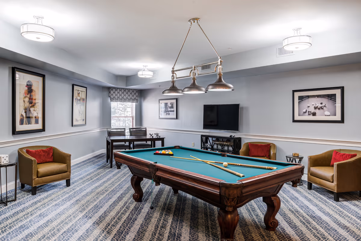 A recreational room with a pool table in the center, surrounded by three brown armchairs with red cushions. The room has patterned carpet flooring, framed artwork on the walls, a flat-screen TV mounted on the wall, and a small table with two chairs near a window with a patterned valance. Overhead lighting fixtures illuminate the space.