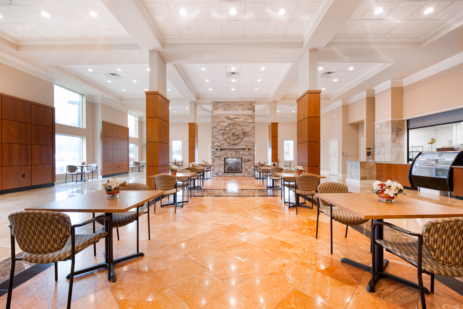 Spacious dining area with multiple tables and chairs arranged neatly. The room features large windows allowing natural light, a stone fireplace with a decorative clock above it, and a counter area with a display case on the right side. The floor is made of polished orange marble tiles, and the ceiling has recessed lighting.