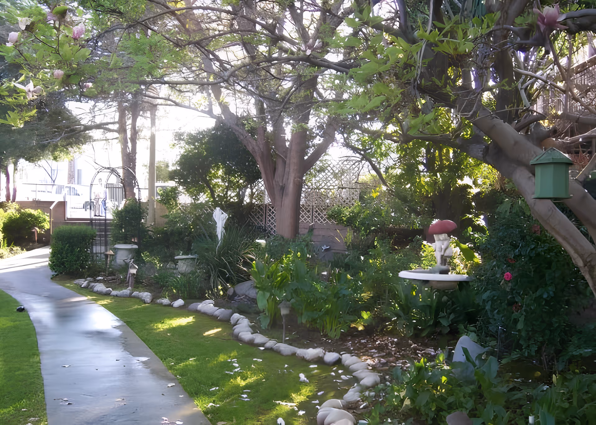 Shaded courtyard garden with a winding paved path, trees, flowering shrubs, stone border, and a birdbath with a small statue.