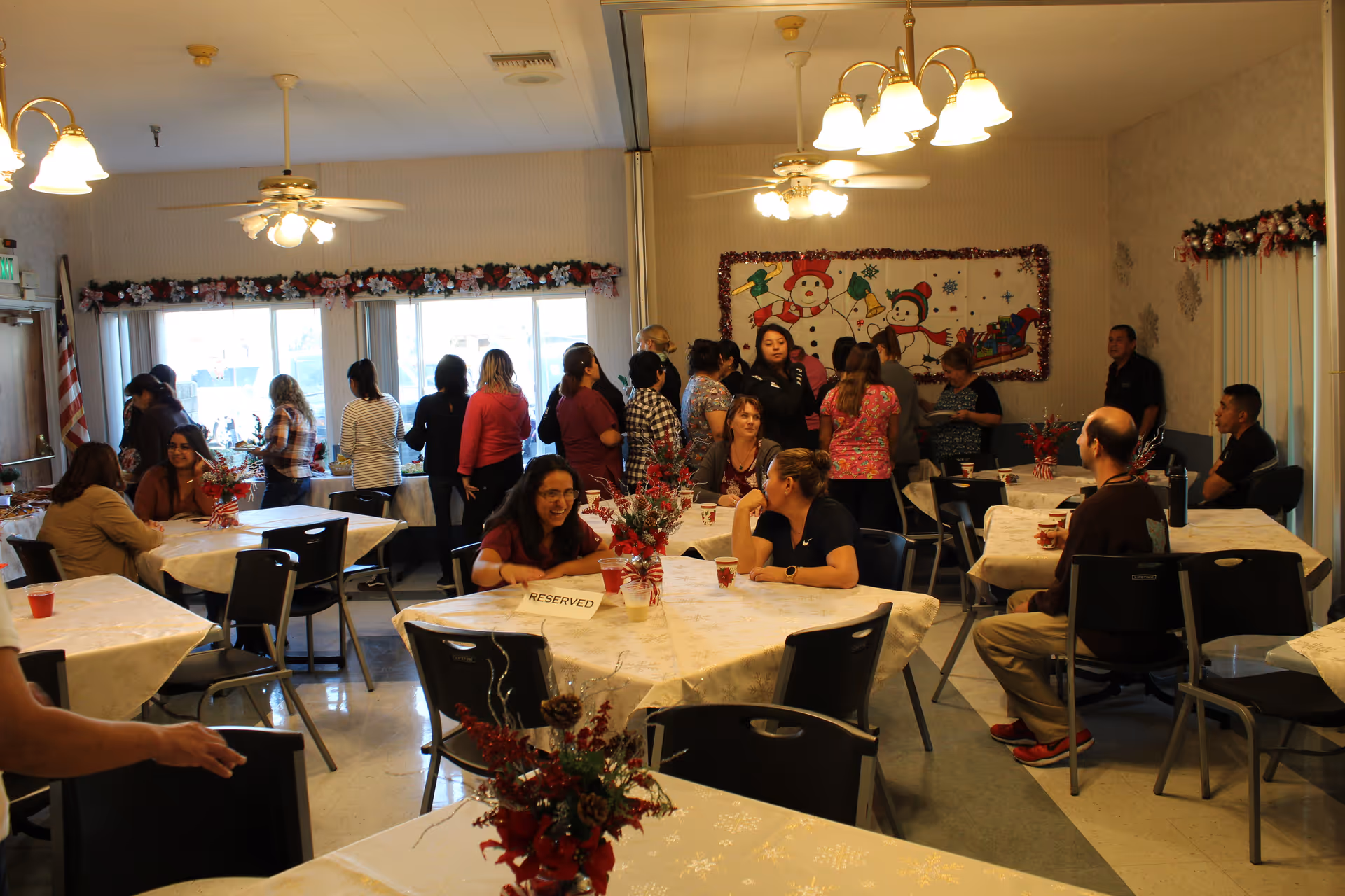 A group of people gathered in a decorated dining room with tables covered in white tablecloths and floral centerpieces. Some people are seated and chatting, while others stand near the windows. The room has ceiling fans with lights and holiday decorations including a wall hanging with snowmen and garlands.