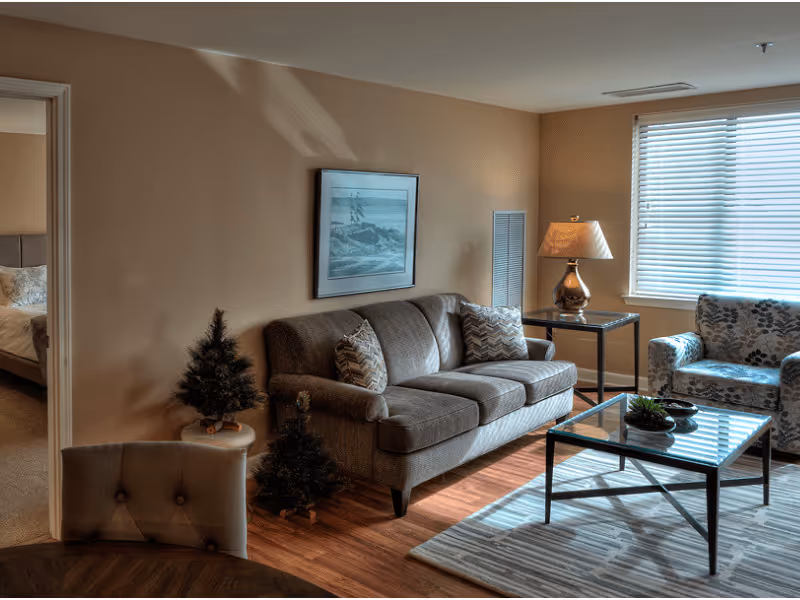 A cozy living room with a gray sofa adorned with patterned cushions, a glass coffee table with a small plant, a floral armchair, and a side table with a lamp. Sunlight streams through a window with blinds, casting shadows on the wooden floor. A framed picture hangs on the beige wall above the sofa. To the left, an open doorway reveals a bedroom with a bed and pillows.