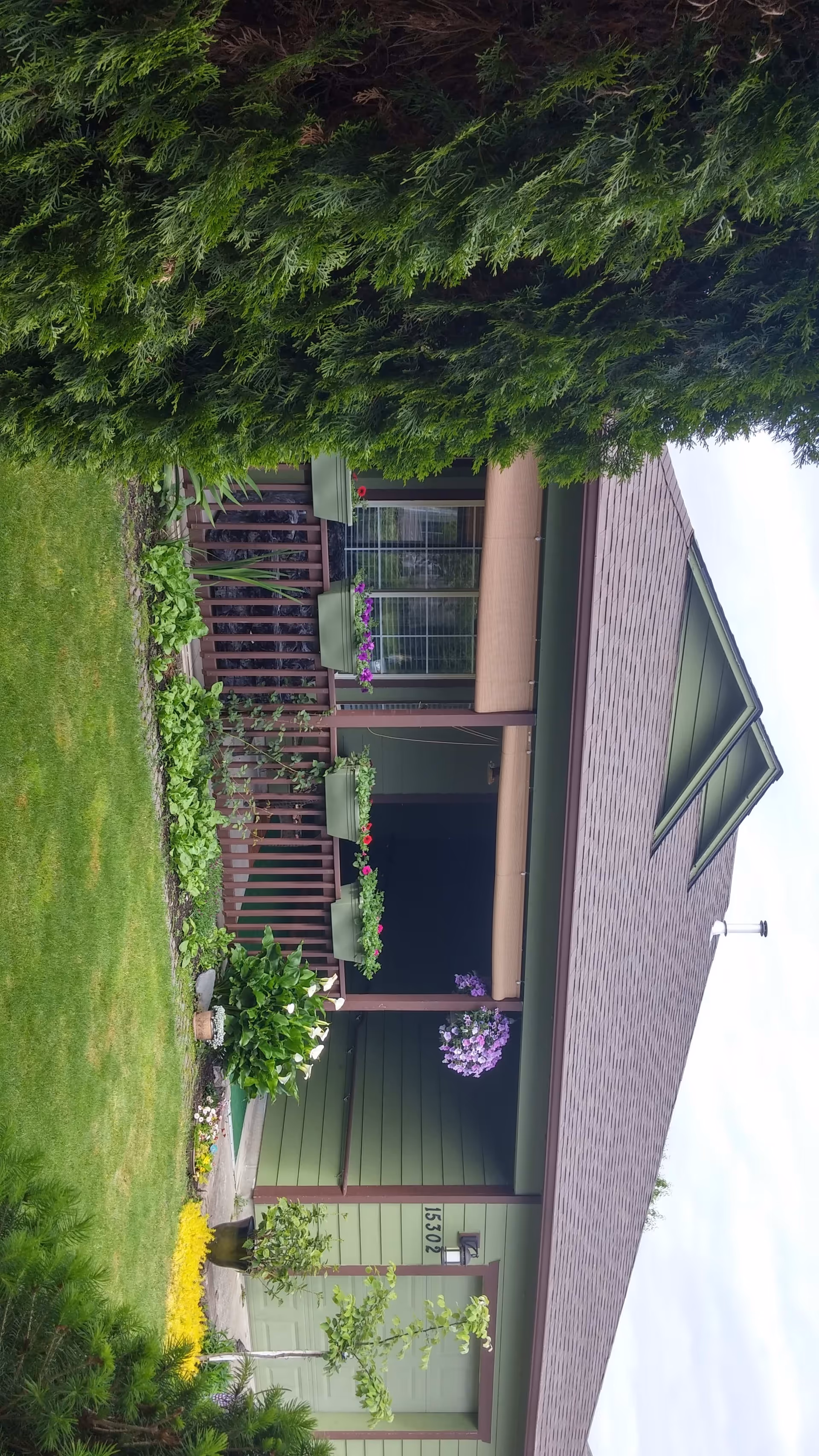 Front exterior view of a green house with a brown roof and a porch decorated with flower pots and hanging plants. The house number 15302 is visible near the garage door. The lawn is well-maintained with various plants and shrubs.