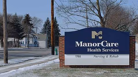 Blue ManorCare Health Services sign on brick base beside a snowy sidewalk and trees