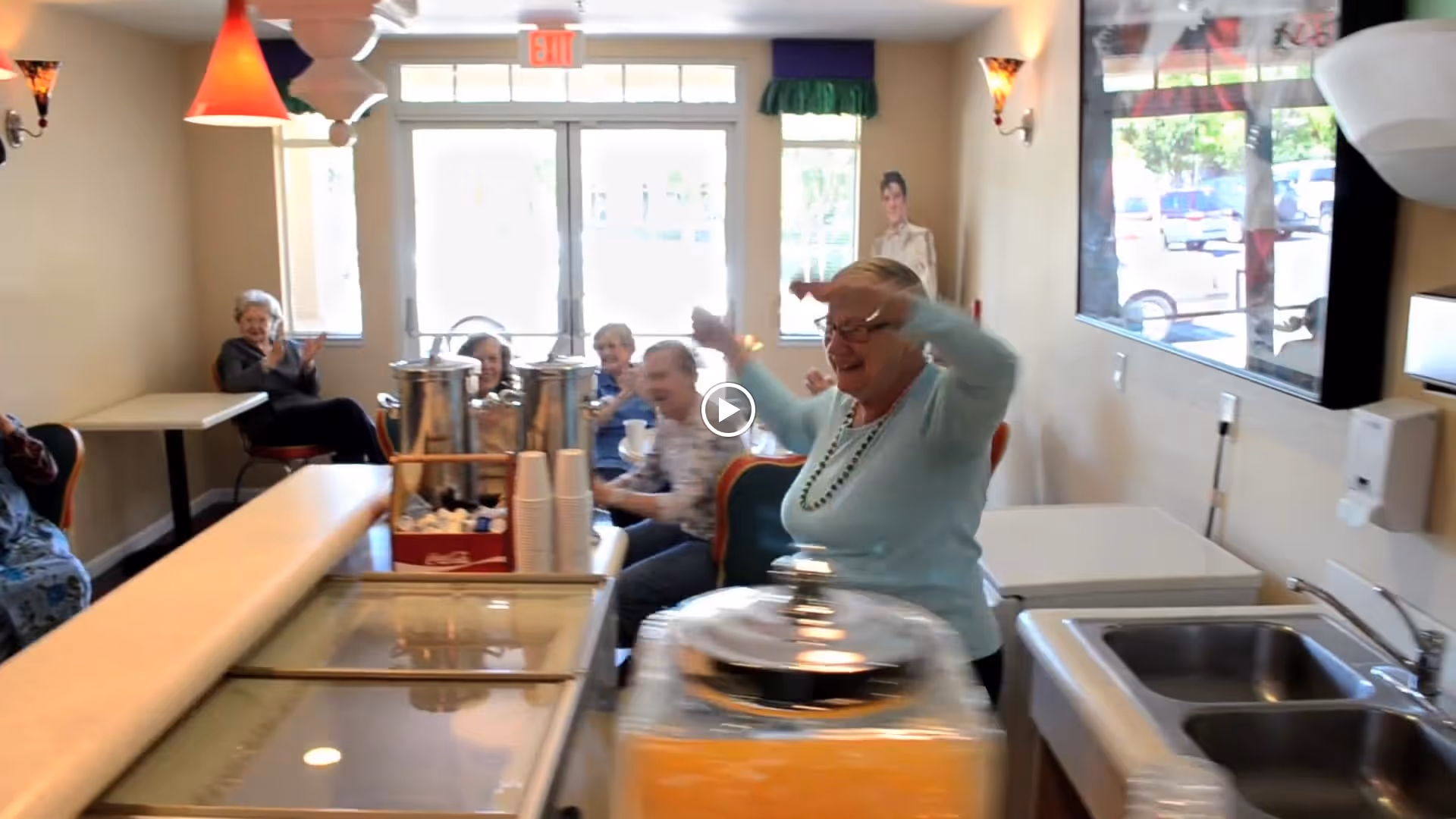 A group of elderly people sitting and clapping in a bright room with large windows. In the foreground, a woman with glasses and a light blue sweater is raising her arms joyfully near a counter with a glass display case and a sink.