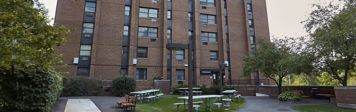 Outdoor courtyard area of The Bernardine facility featuring picnic tables, benches, green grass, trees, and a tall brick building in the background.