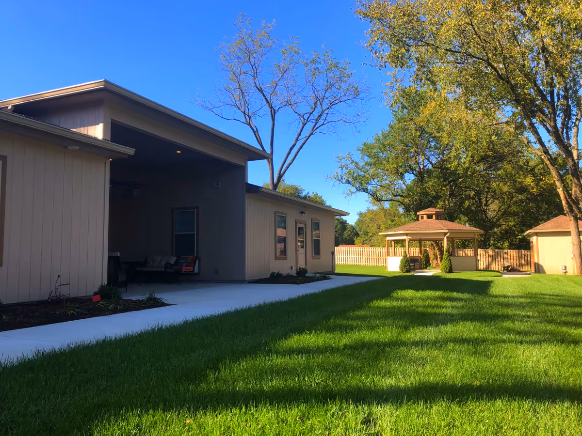 Outdoor view of a senior care facility showing a beige building with a covered patio area furnished with chairs, a concrete walkway, green grass lawn, trees, and a wooden gazebo with seating in the background under a clear blue sky.