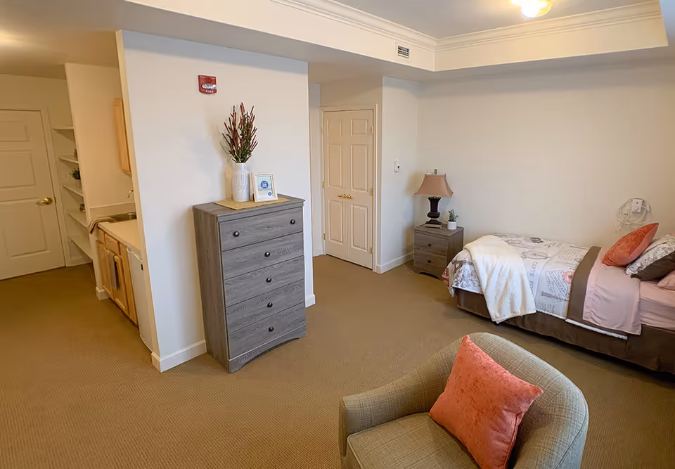 A cozy assisted living bedroom featuring a single bed with patterned bedding and pillows, a wooden nightstand with a lamp and small plant, a gray chest of drawers with a vase of flowers and a framed picture on top, a comfortable armchair with a coral cushion, and a small kitchenette area with a sink and cabinets in the background.