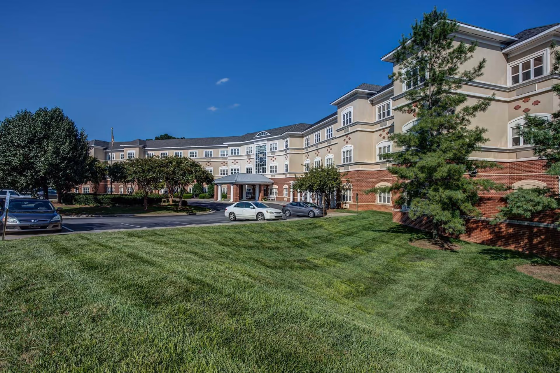 Exterior view of a large senior living facility building with beige and red brick walls, multiple windows, and a covered entrance. There is a well-maintained green lawn in the foreground, several trees, and a few parked cars near the entrance under a clear blue sky.