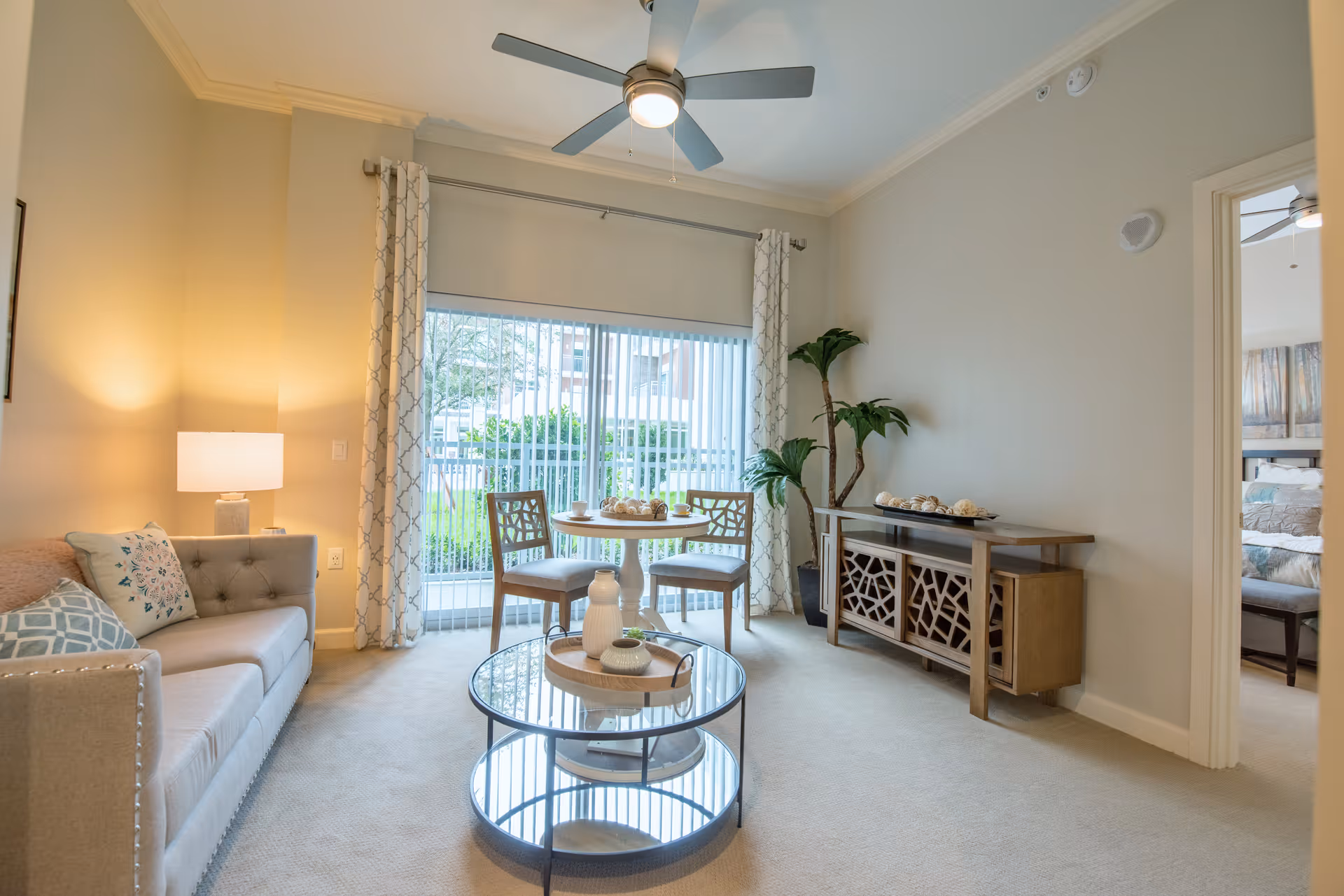 A bright living room with a beige sofa adorned with patterned pillows, a round glass coffee table with decorative items, a small round dining table with two chairs near a large window with vertical blinds and patterned curtains, a ceiling fan with light, a decorative wooden console table with a plant beside it, and an open doorway leading to a bedroom.
