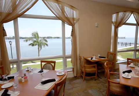 Dining area with wooden tables and chairs set with plates, cups, and black napkins. Large windows with sheer curtains offer a view of a waterfront with palm trees and a pier.