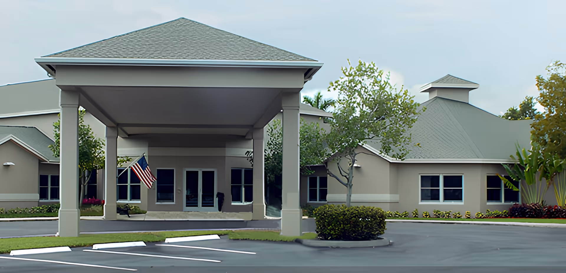 Exterior view of Majestic Memory Care facility showing the entrance with a covered drop-off area, an American flag, surrounding windows, and landscaped greenery including bushes and trees.
