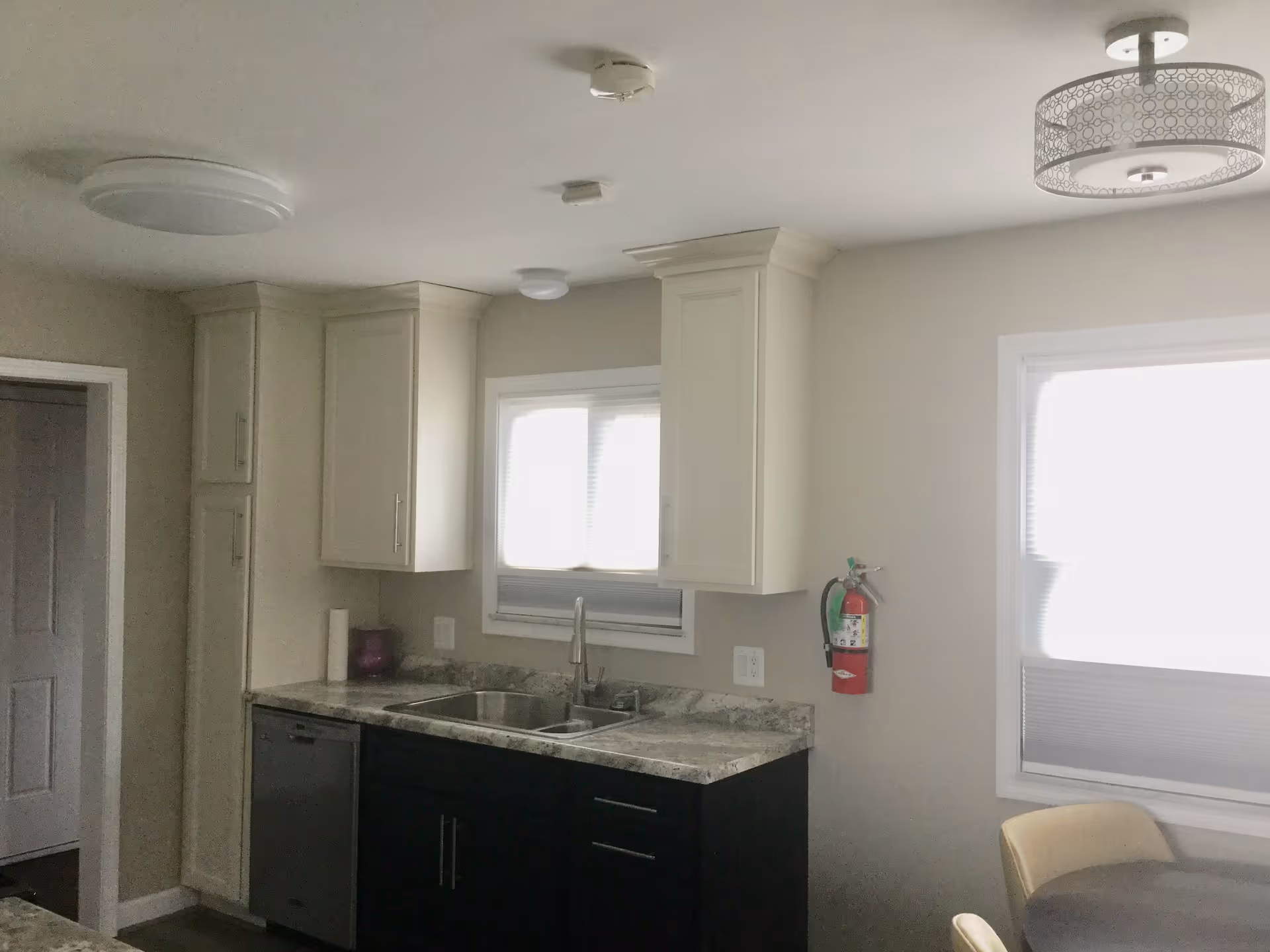 Interior view of a kitchen area with white upper cabinets and dark lower cabinets, a double sink under a window with blinds, a dishwasher, a fire extinguisher mounted on the wall, and a round table with chairs near another window.