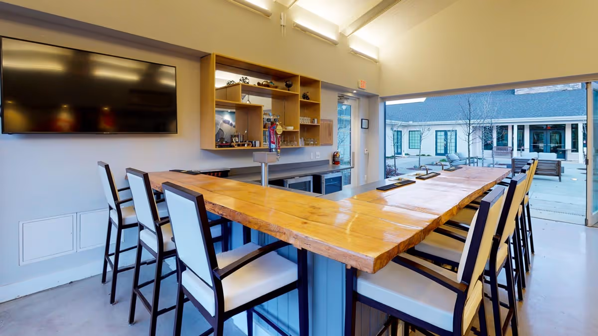 Interior communal bar area with a long wooden countertop surrounded by high-backed chairs, a wall-mounted TV, and large open windows to an outdoor courtyard.