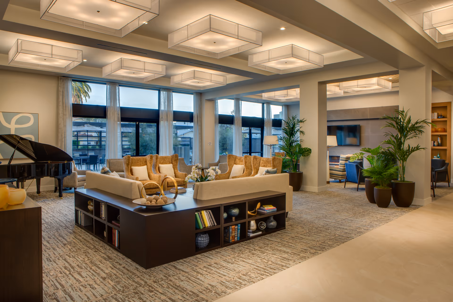 A spacious and well-lit living room area with large windows allowing natural light to fill the space. The room features a central seating arrangement with beige and brown cushioned chairs and sofas around a dark wood coffee table with built-in shelves holding books and decorative items. There are several potted plants adding greenery, modern rectangular ceiling lights, a grand piano in the corner, and a separate seating area with a television visible in the background.
