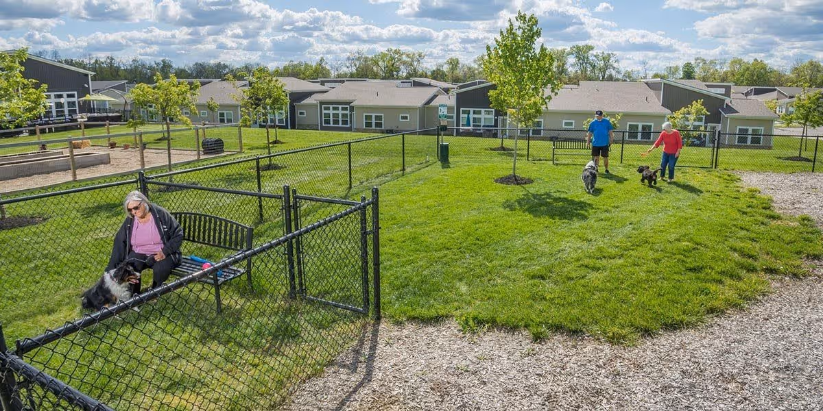 Outdoor fenced dog park area at a senior living facility with green grass, small trees, and a bench. An elderly woman sits on the bench petting a dog inside the fenced area, while an elderly man and woman walk two dogs on the grass outside the fenced area. Residential buildings are visible in the background under a partly cloudy sky.
