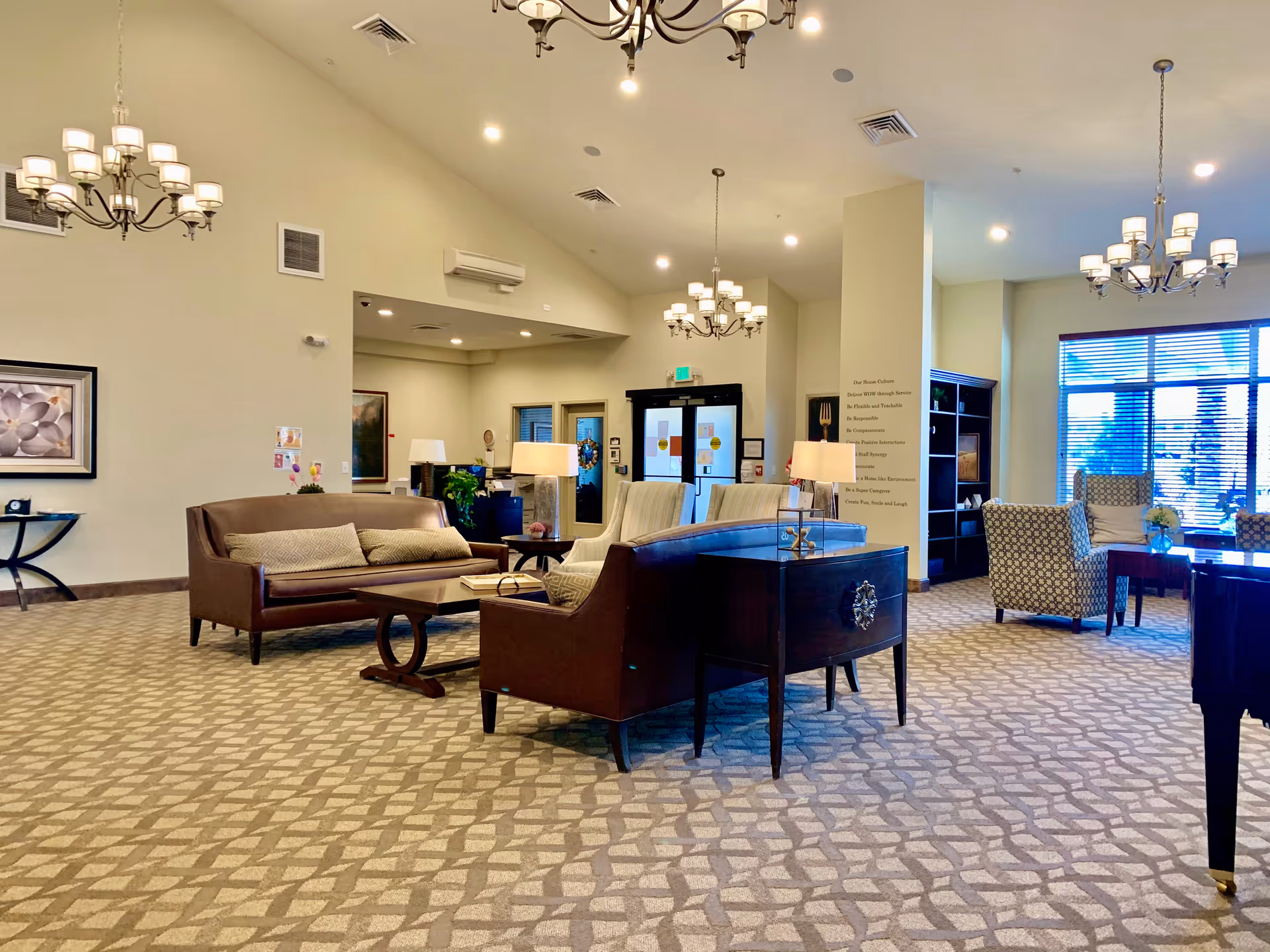 Spacious, well-lit assisted living facility lounge with sofas, armchairs, chandeliers, and large windows.