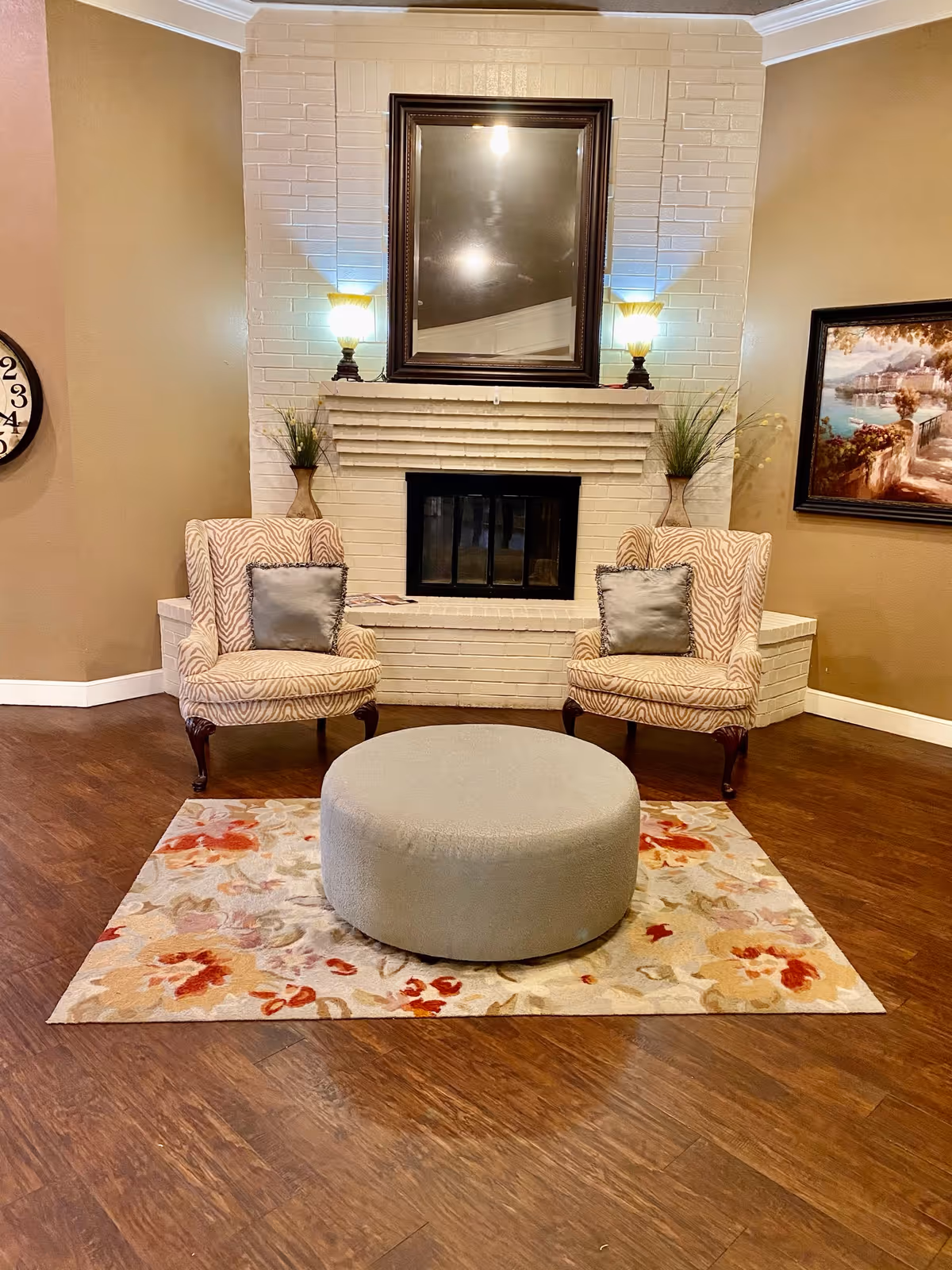 A cozy sitting area with two patterned armchairs each with a gray pillow, positioned on either side of a white brick fireplace. Above the fireplace is a large mirror flanked by two lamps. In front of the chairs is a round gray ottoman on a floral area rug. The room has wooden flooring and beige walls, with a framed painting on the right wall and a clock partially visible on the left wall.