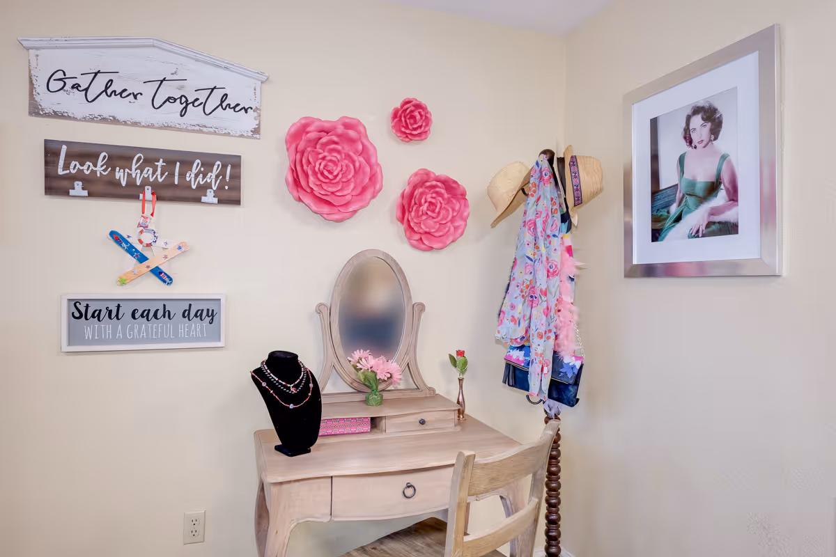 A cozy corner with a wooden vanity table and chair. The vanity has a small mirror, a vase with pink flowers, and a black jewelry display with necklaces. The wall above the vanity features three decorative signs with the phrases 'Gather Together,' 'Look what I did!' and 'Start each day with a grateful heart.' There are also three large pink flower wall decorations and a coat rack holding hats and colorful scarves. A framed portrait of a woman in a green dress hangs on the adjacent wall.