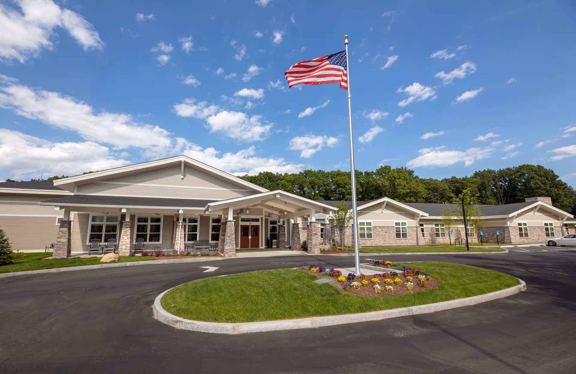 Exterior view of a single-story senior living facility building with a covered entrance, stone and siding facade, and an American flag on a flagpole in the center of a circular driveway. The sky is partly cloudy and there are trees in the background.