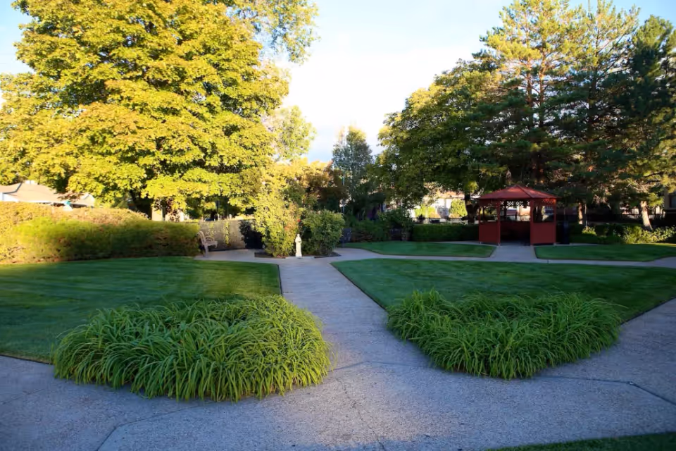 A well-maintained outdoor garden area with green grass, bushes, and large trees. There are paved walkways leading to a red gazebo in the background, surrounded by more greenery and trees.