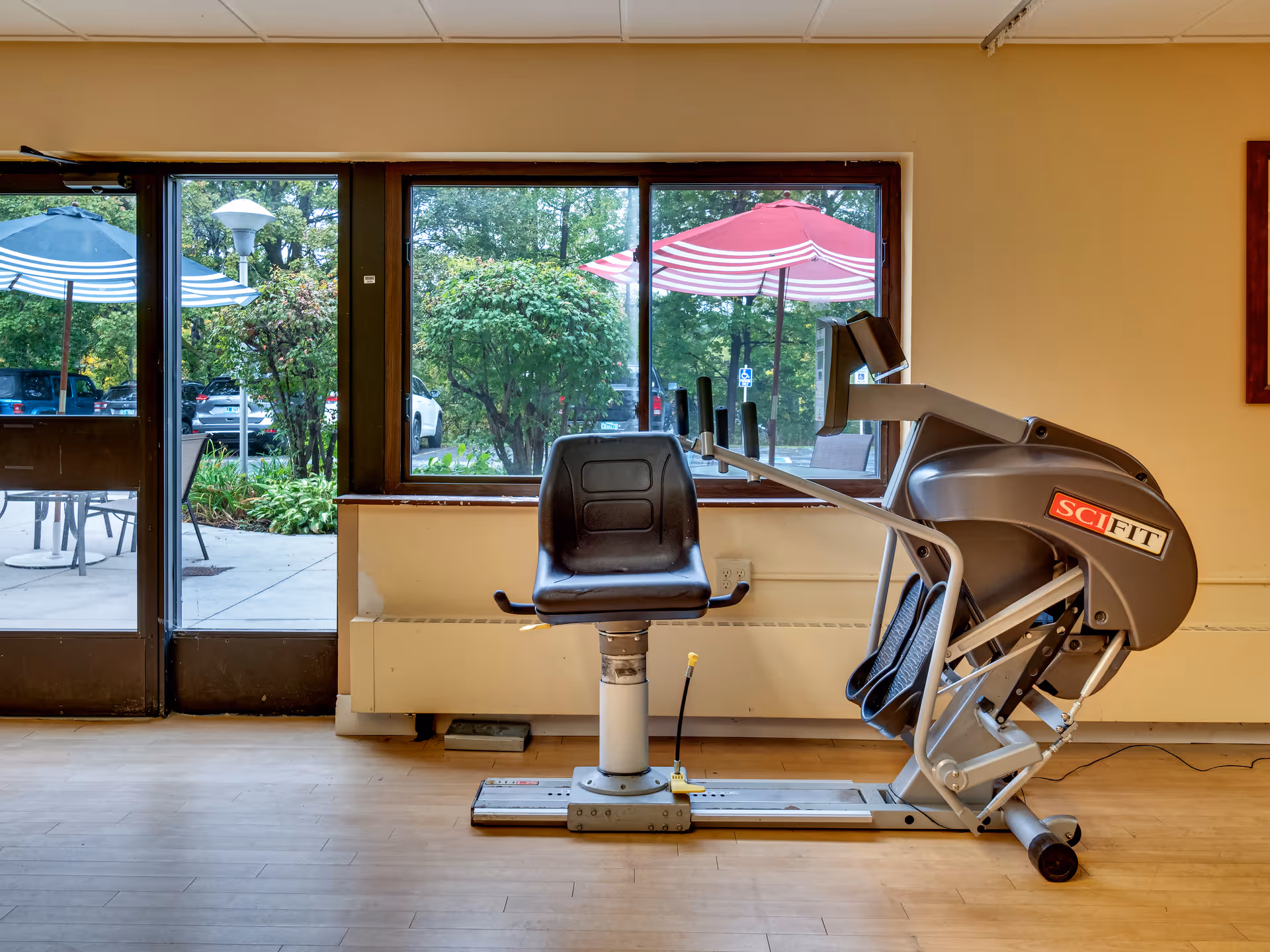 A seated exercise machine with a black seat and foot pedals positioned in front of a large window. Outside the window, there are patio tables with umbrellas and greenery. The room has light-colored walls and wooden flooring.