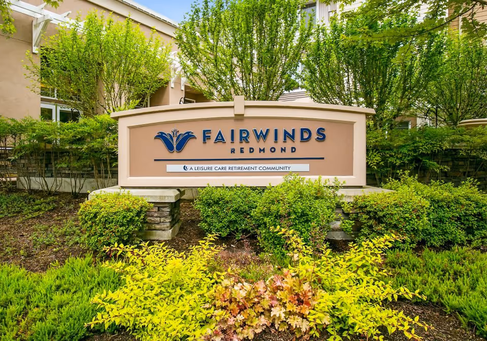 Outdoor view of the entrance sign for Fairwinds Redmond, a leisure care retirement community, surrounded by green bushes and trees with part of the building visible in the background.