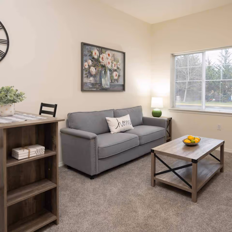 A cozy living room with a gray sofa adorned with a white pillow that says 'home', a wooden coffee table with a bowl of fruit, a side table with a green lamp, a large window with blinds, a wall clock, and a painting of white flowers in a vase. The room has beige walls and carpeted flooring.
