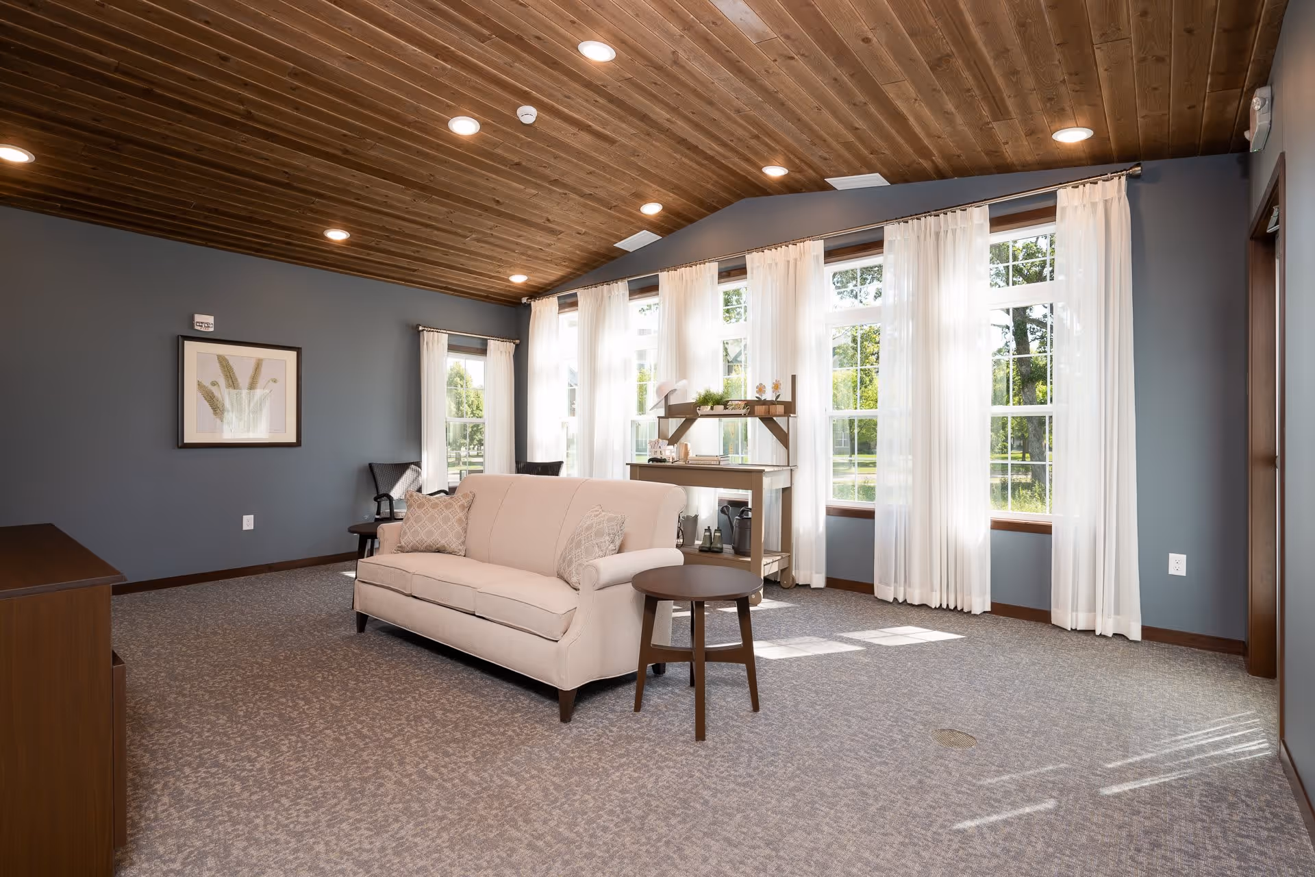 Bright senior living lounge with a beige sofa, round side table, large windows with white curtains, and a wood-paneled ceiling.