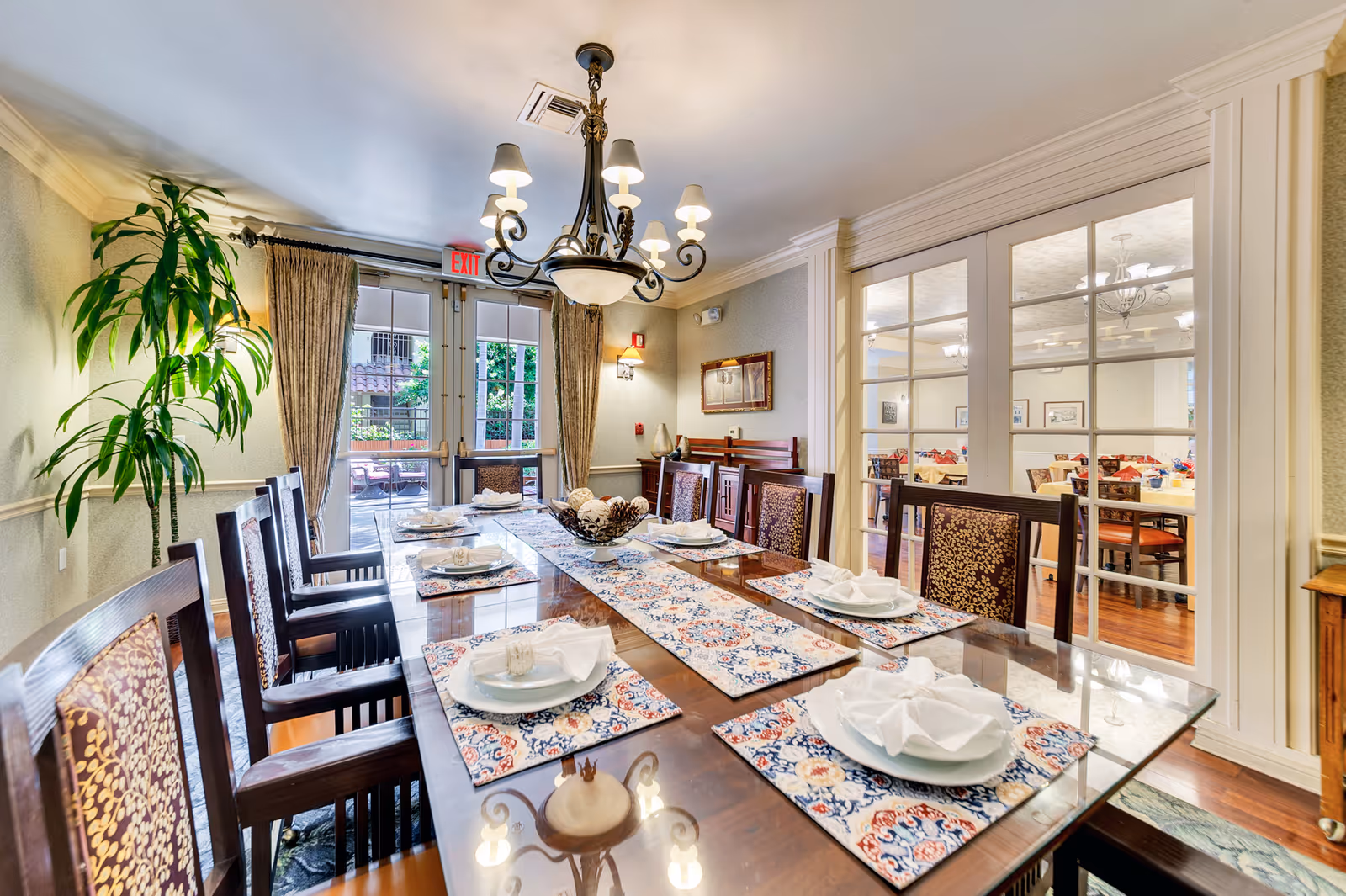 A formal dining room with a long set table, upholstered chairs, a chandelier, and glass doors leading to another seating area and an outdoor patio.