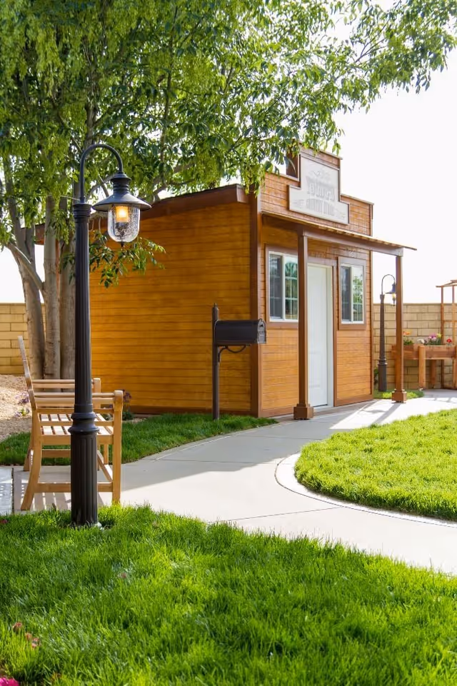 A small wooden building with a white door and two windows, situated in a garden area with green grass, a curved concrete pathway, a wooden bench, and black vintage-style lamp posts. Trees and a brick wall are visible in the background.