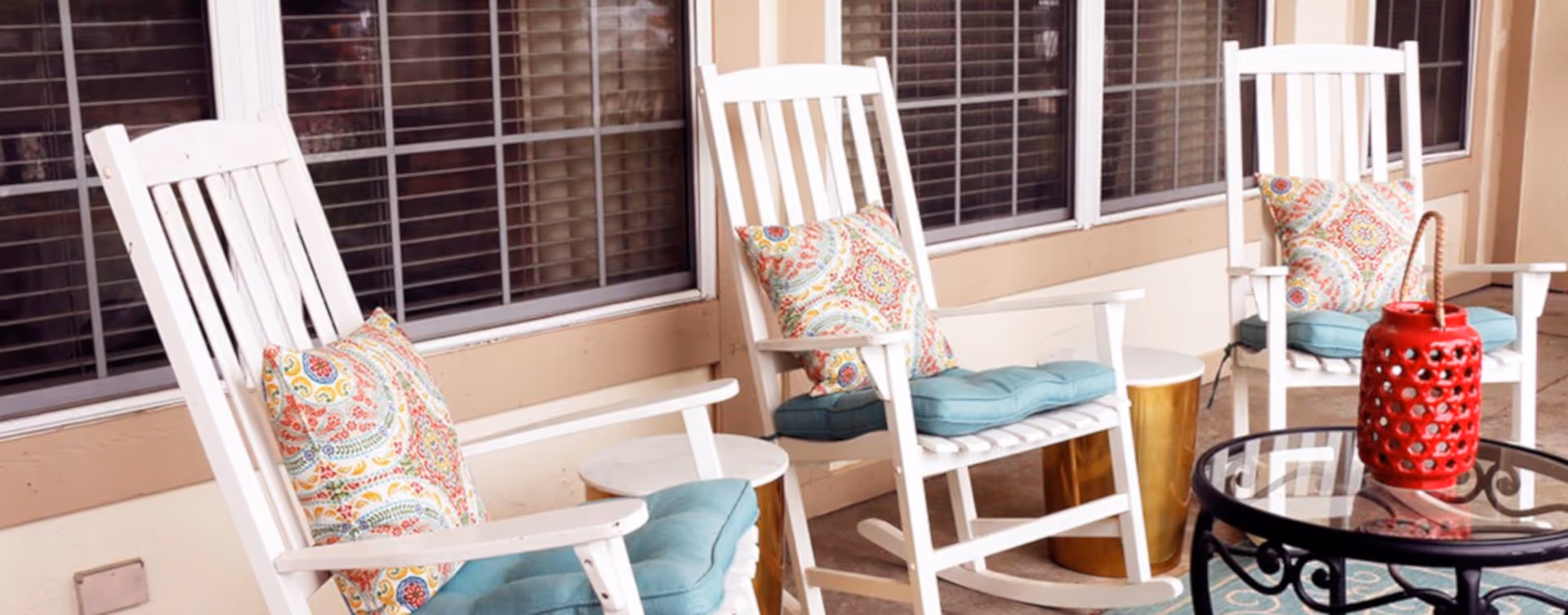 A covered porch with white rocking chairs topped with teal seat cushions and patterned pillows around small tables and a glass-top table with a red decorative lantern.