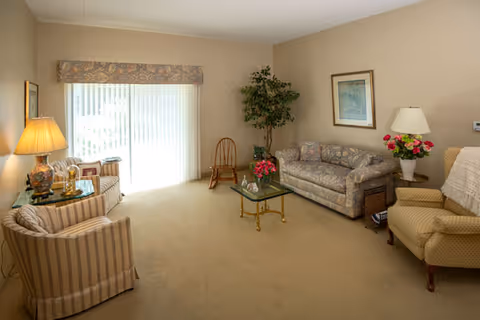 A cozy living room with beige walls and carpet, featuring a striped armchair with a lamp on a glass side table, a floral patterned sofa, a glass coffee table with a flower arrangement, a wooden chair, a potted plant, a framed picture on the wall, and a sliding glass door with vertical blinds letting in natural light.